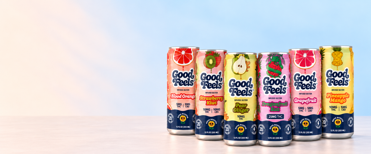 Six Good Feels sparkling beverage cans in different fruit flavors lined up on a light surface against a soft blue gradient background. The cans feature colorful labels with illustrations of citrus, pear, strawberry, grapefruit, and pineapple, and navy blue branding at the bottom.